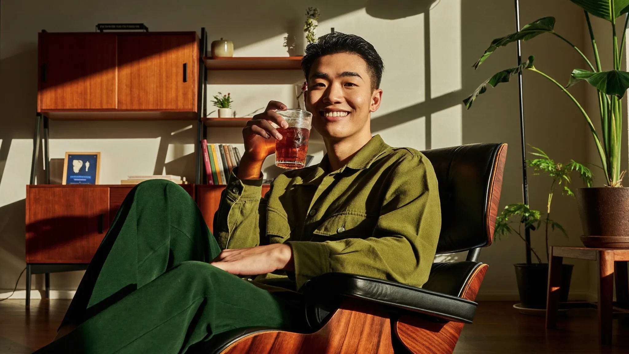 Man in an olive camp-collar shirt relaxing in a walnut mid-century lounge chair in a sunlit living room